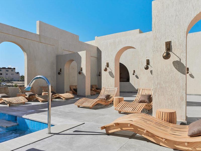 Moderner Poolbereich mit geschwungenen Holzliegen, hellen Wänden und klarem blauem Himmel.