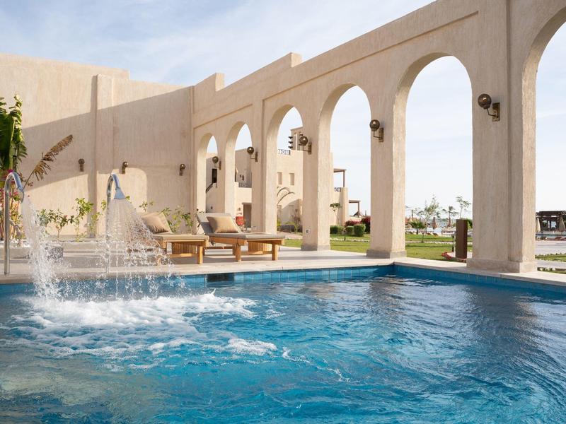 Piscina esterna con fontana accanto ad archi e lettini sotto un cielo soleggiato.