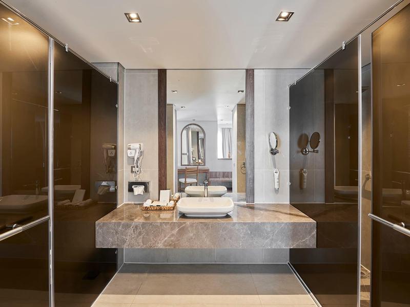 Moderno baño de hotel con gran espejo, lavabo doble y elegantes armarios marrones en las paredes.