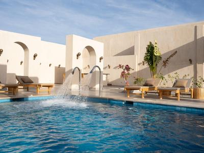 Piscina blu con getti d'acqua e lettini davanti a pareti beige sotto un cielo azzurro.