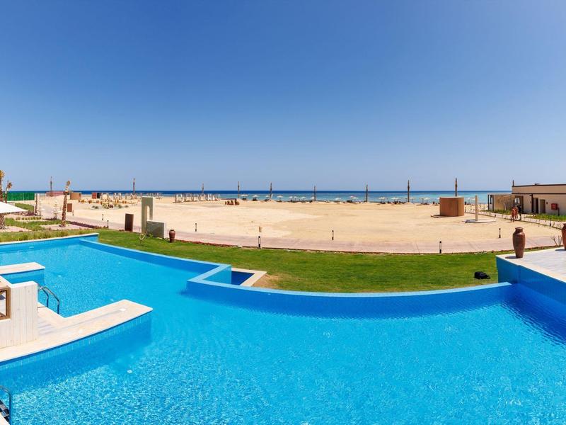 Otwarty basen z leżakami przy piaszczystej plaży pod jasnym błękitnym niebem.