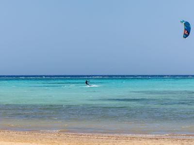 Person kitesurft auf türkisfarbenem Meer bei klarem Himmel am Sandstrand.