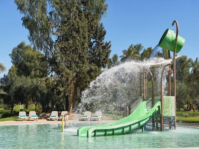 Wasserpark mit Rutsche und Wassereimer, umgeben von Bäumen und Liegestühlen.