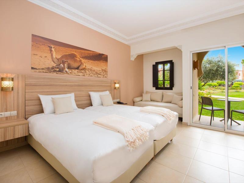 Světý hotelový pokoj se dvěma postelemi, křeslem a výhledem na terasu a zahradu.