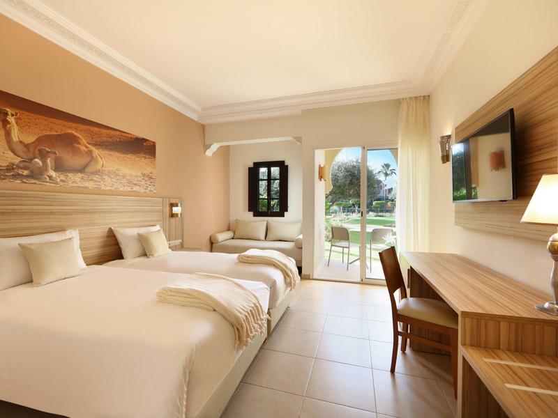 Světle hotelový pokoj se třemi postelemi, stolem, televizí a výhledem na zahradu.