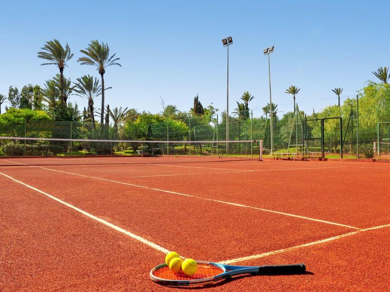 Venku červený antukový tenisový kurt s raketou a míčky v popředí za slunečného dne.