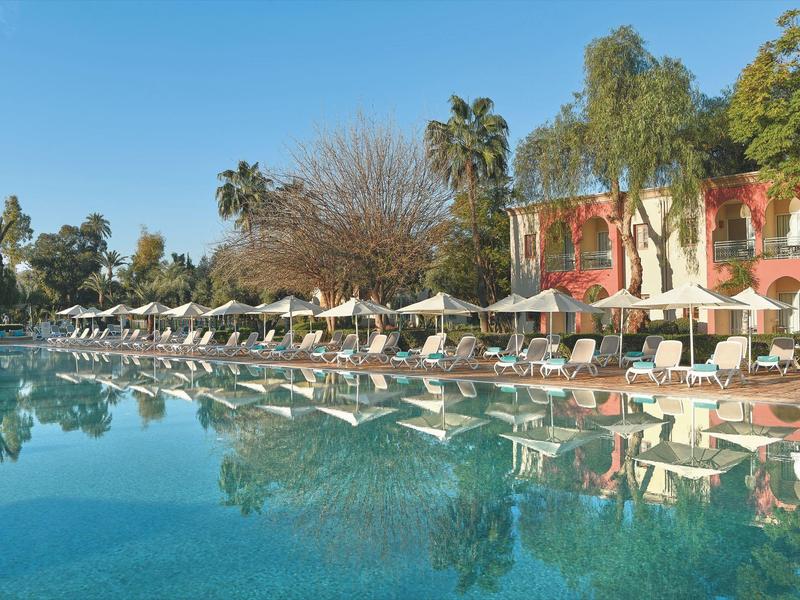 Velký bazén před hotelem s lehátky a slunečníky, obklopený stromy a palmami.