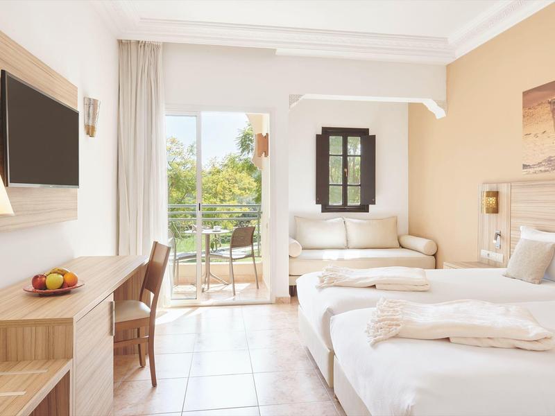 Světý hotelový pokoj se dvěma postelemi, psacím stolem, televizí a balkonem s výhledem na zeleň.