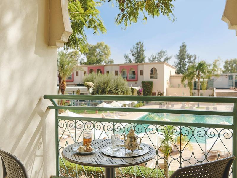 Balkón s stolem a židlemi s výhledem na bazén a hotelové budovy za denního světla.