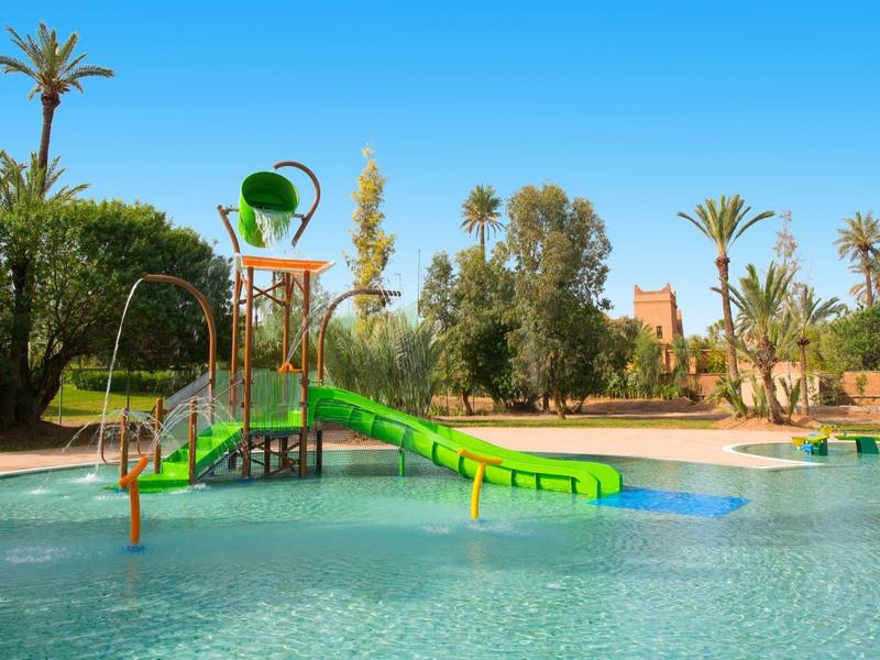 Vodní hřiště s tobogánem a kbelíkem v bazénu obklopeném palmami pod jasně modrou oblohou.