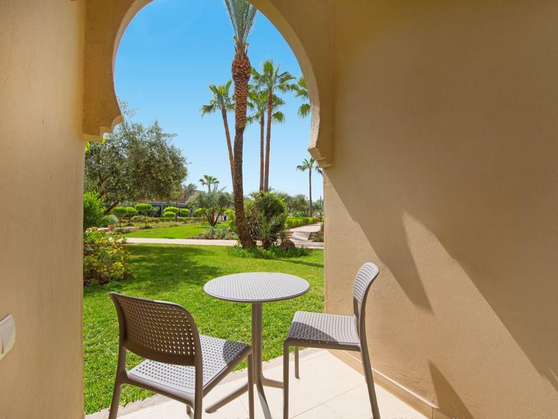 Balkon se dvěma židlemi a malým stolem s výhledem na upravenou zahradu a palmy.