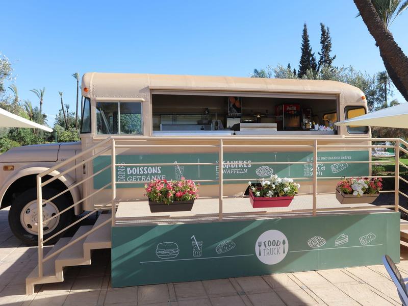 Přestavěný autobus jako kavárna s posezením a rostlinami na terase za slunečného počasí.