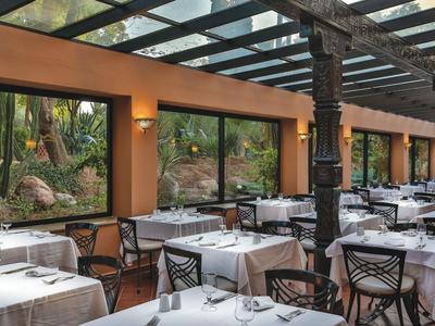 Elegant restaurant with set tables and large windows overlooking tropical plants.