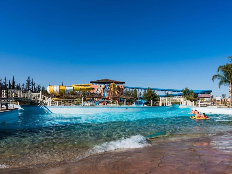 Grand piscine d'hôtel avec toboggans aquatiques, parasols et palmiers sous un ciel bleu clair.