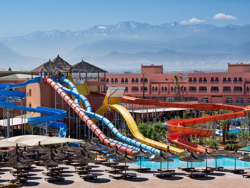 Parc aquatique coloré avec toboggans devant un complexe hôtelier et montagnes en arrière-plan.