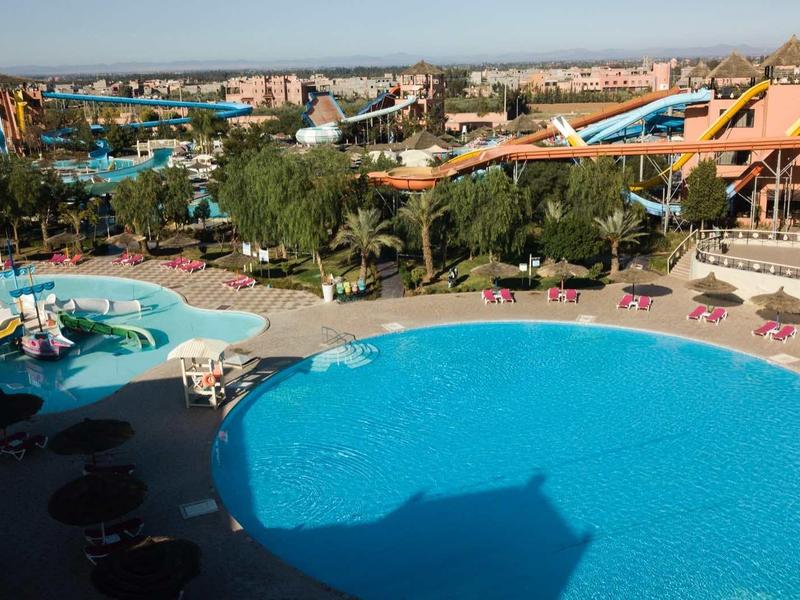 Grand piscine extérieure avec parasols, entourée de bâtiments et parc aquatique au loin.