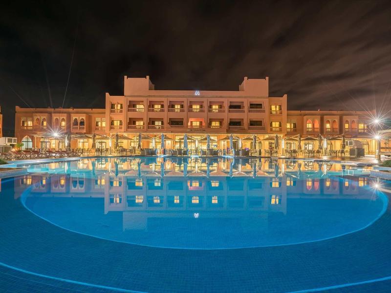 Vue nocturne d'un hôtel illuminé avec une grande piscine calme au premier plan.