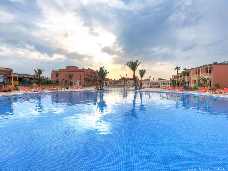 Grand piscine d'hôtel au coucher du soleil avec des palmiers et des bâtiments rouges en arrière-plan.