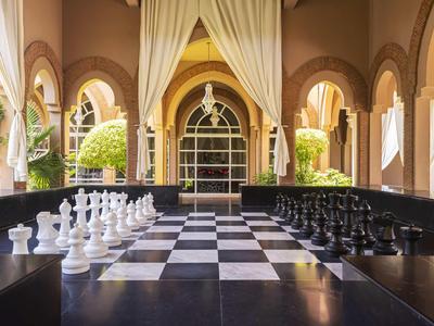 Velká šachovnice na hotelové terase s obloukovými okny a rostlinami.