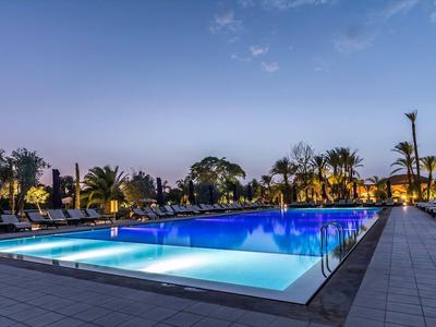 Abendliche Aufnahme eines beleuchteten Pools mit Liegestühlen und Palmen im Hintergrund.