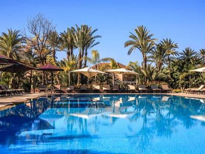 Klares Schwimmbecken mit Liegestühlen, Sonnenschirmen und Palmen unter blauem Himmel.