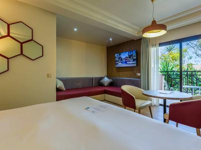 Moderní hotelový pokoj s postelí, pohovkou, televizí a balkonem s výhledem na zelené stromy.