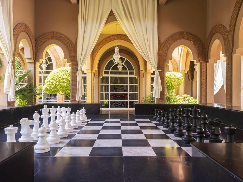 Velká šachovnice na hotelové terase s obloukovými okny a rostlinami.