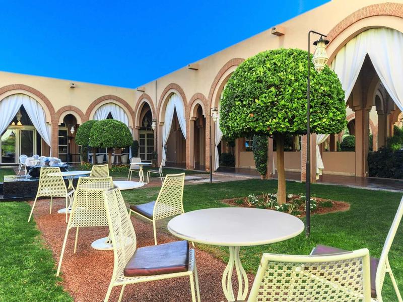 Dvůr s kulatými stoly, židlemi a upravenými stromy před budovou s oblouky a závěsy.