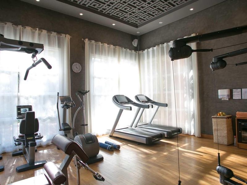 Prosvětlená posilovna s běžeckými pásy a posilovacími stroji u velkých oken s bílými záclonami.