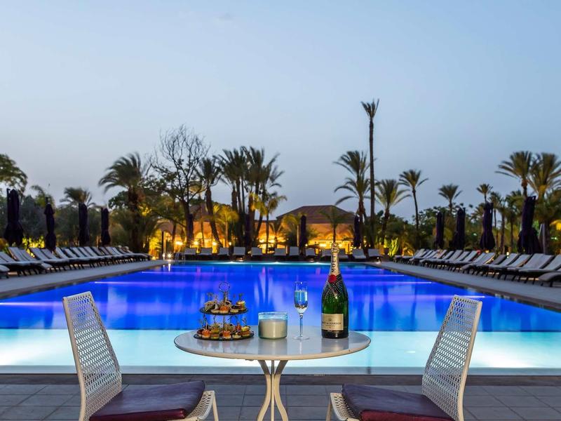 Osvětlený hotelový bazén s dvěma židlemi a šampaňským na stole v popředí za soumraku.