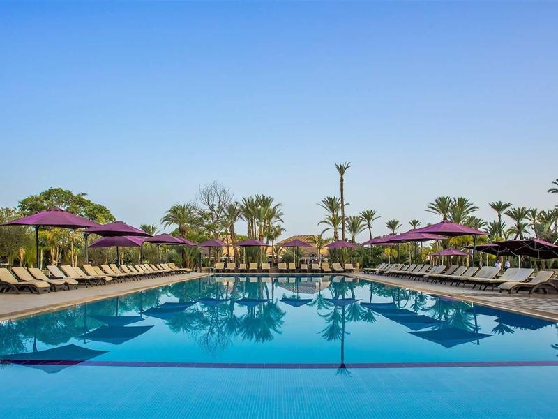 Velký venkovní bazén s lehátky a slunečníky pod jasnou oblohou.