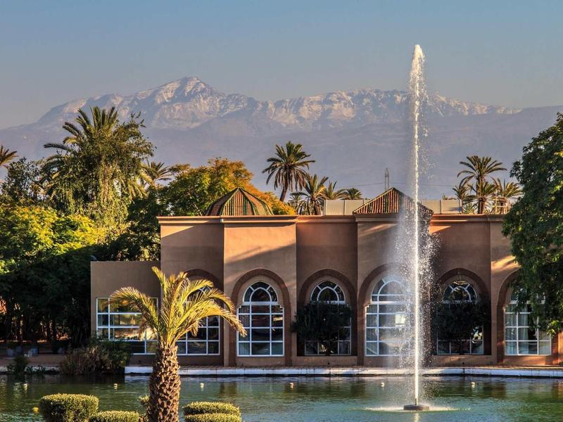 Hotelová budova s obloukovými okny, hory v pozadí a vodotrysk v zahradě.