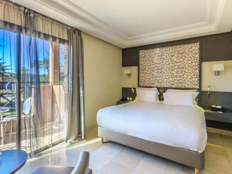 Světý hotelový pokoj s velkou postelí, denním světlem a výhledem na terasu s palmami.