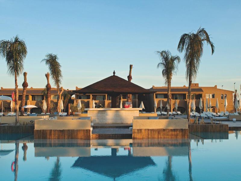 Hotel with pool, sun loungers, and palm trees at sunset under clear sky.