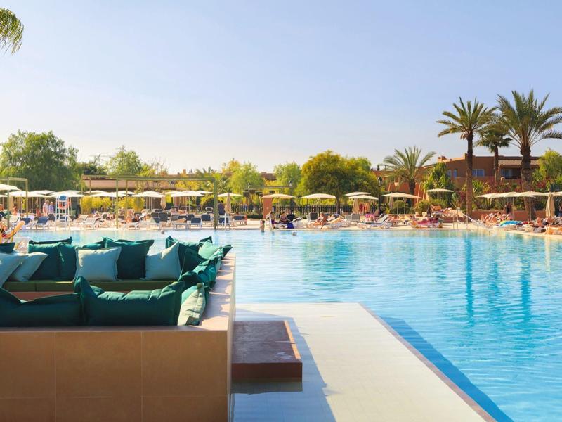 Large outdoor pool with loungers and umbrellas surrounded by palm trees and blue sky.