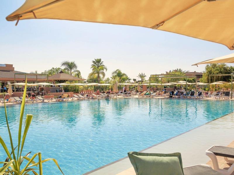 Large outdoor pool with umbrellas and lounge chairs, surrounded by palm trees and buildings.