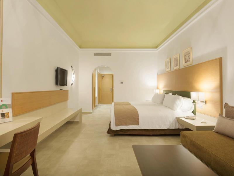 Modern hotel room with large bed, desk, and seating area in neutral tones.