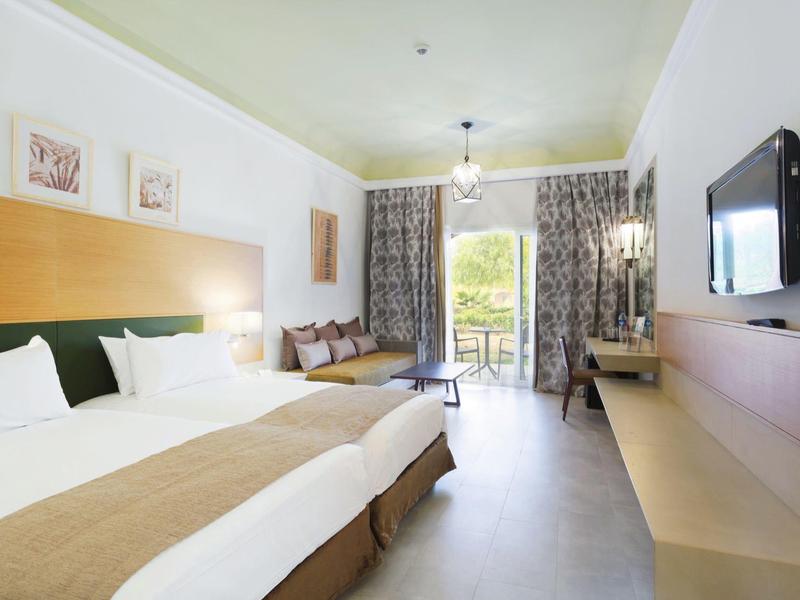 Bright hotel room with two beds, balcony, and modern wall-mounted TV.