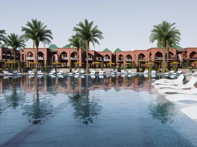 Großer Pool mit weißen Liegestühlen und Palmen vor einem Resortgebäude bei klarem Himmel.