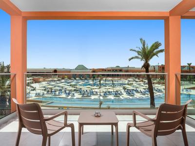 Balcon avec deux chaises et table donnant sur la piscine de l'hôtel et des palmiers sous un ciel clair.