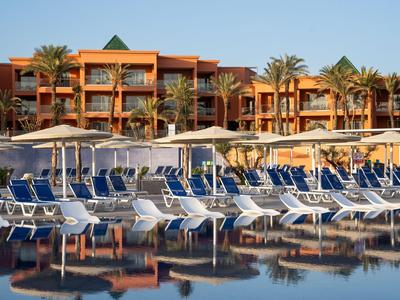 Hôtel à plusieurs étages avec piscine et rangées de chaises longues bleues sous parasols blancs.