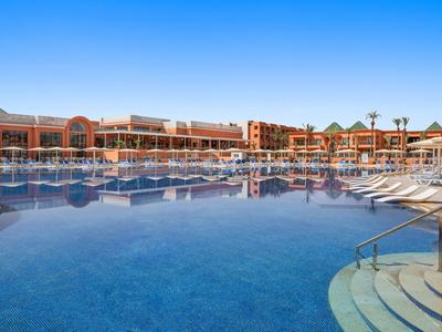 Grande piscine extérieure avec chaises longues devant un bâtiment hôtelier sous un ciel bleu clair.