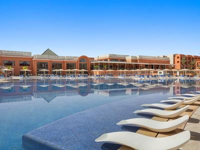 Grand hôtel avec piscine extérieure et transats sous un ciel bleu clair.