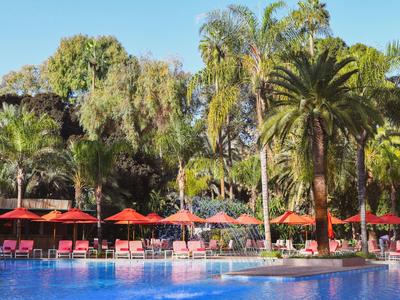Großer blauer Swimmingpool mit roten Sonnenschirmen und Palmen im Hintergrund bei sonnigem Wetter.