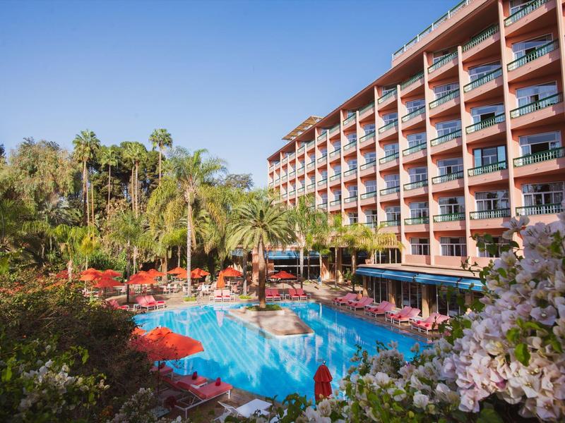 Budynek hotelu z basenem, palmami i czerwonymi leżakami w słonecznym otoczeniu.