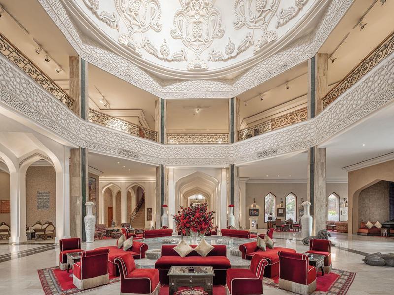 Grande hall d'hôtel avec plafonds ornés et canapés en velours rouge disposés au centre.