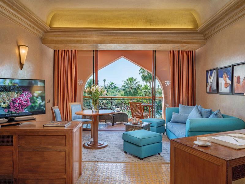 Salon lumineux avec canapé bleu, télévision, meubles en bois et balcon donnant sur des palmiers.