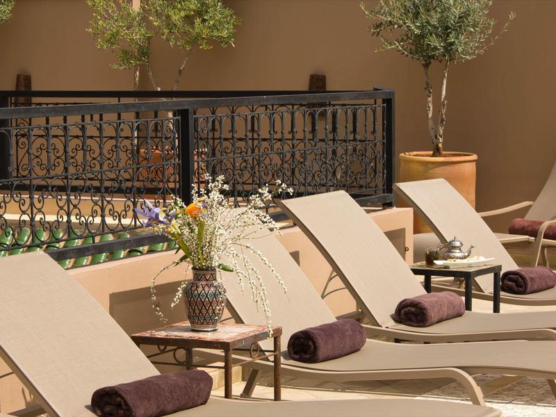 Multiple light lounge chairs with dark towels by a hotel pool with tables and flowers.