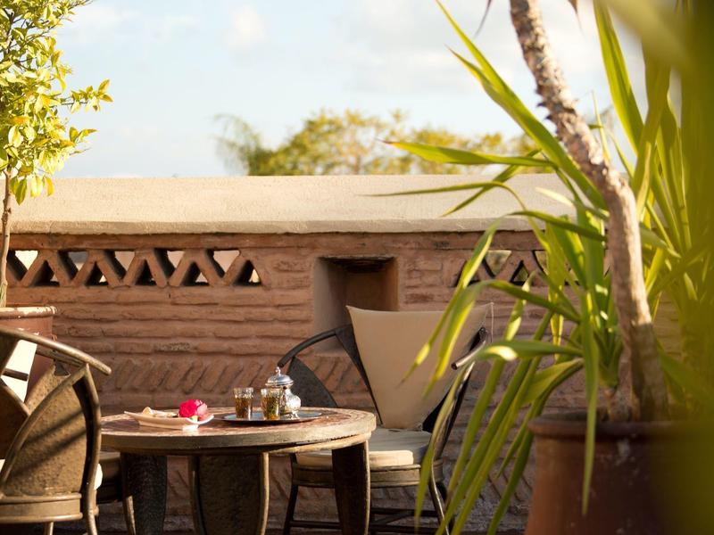 Sunny terrace with wooden tables, chairs, and plants in front of a brick wall.