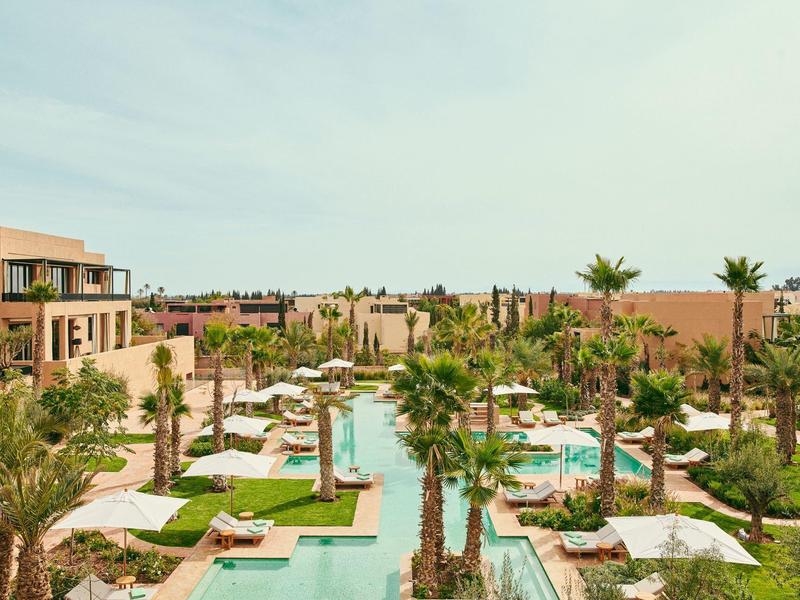 Aerial view of a resort with pool, palm trees, and beige buildings under a blue sky.